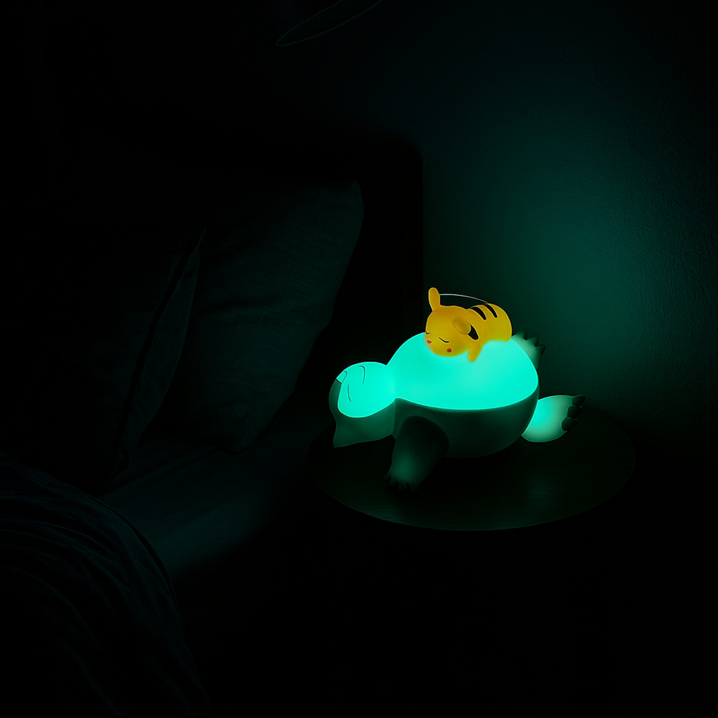 Lampe LED sans fil en forme de Ronflex avec Pikachu endormi sur son ventre, éclairée dans une chambre sombre sur une table de chevet. Idéal comme veilleuse Pokémon pour enfant ou décoration geek.
